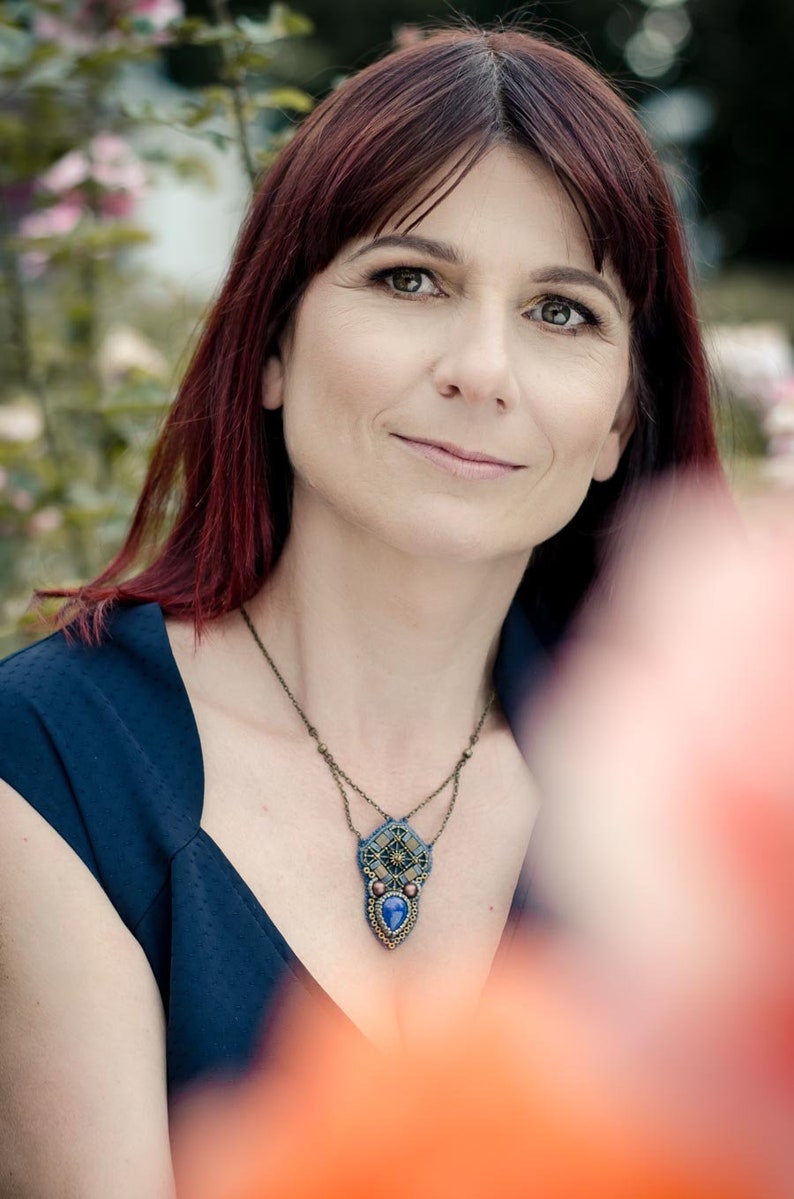 Puede incluir: Una mujer con el pelo largo y rojo que lleva una blusa azul oscuro sin mangas con un collar de cuentas con un colgante de piedra azul.
