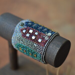 May include: A colorful beaded bracelet with a silver bicycle charm. The bracelet is made with a variety of beads, including iridescent, silver, and gold beads. The bracelet is displayed on a dark wooden stand.