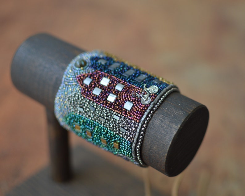 May include: A colorful beaded bracelet with a silver bicycle charm. The bracelet is made with a variety of beads, including iridescent, silver, and gold beads. The bracelet is displayed on a dark wooden stand.