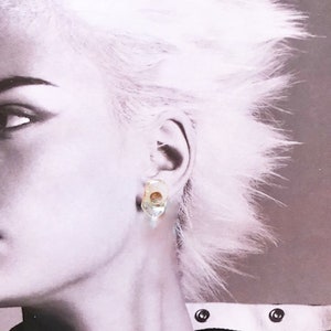 May include: A close-up of a single earring. The earring has a clear, irregular shape with a small, round, brown accent. The earring is set against a black and white background featuring a person's face and hair.