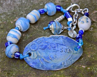 Könnte beinhalten: Ein blaues und weißes Keramikarmband mit einem großen, ovalen Anhänger. Der Anhänger hat eine strukturierte Oberfläche und ist mit einem wirbelnden Muster verziert. Das Armband besteht aus einer Vielzahl von blauen und weißen Perlen, darunter einige mit einem Wirbelmuster.