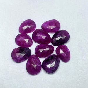 Pode incluir: Uma coleção de nove pedras preciosas facetadas em forma de gota, de cor magenta profunda. As gemas têm facetas mais claras que refletem a luz. As pedras estão dispostas sobre uma superfície branca.