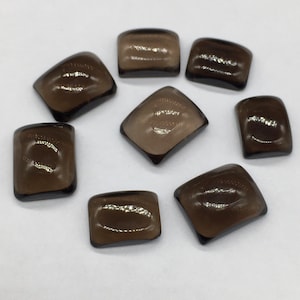 May include: Eight smoky quartz cabochons, each with a slightly different shape and size, are arranged on a white surface. The stones are translucent and have a dark brown colour.