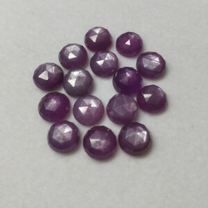 K&ouml;nnte beinhalten: Eine Gruppe von 15 violetten facettierten Edelsteinen, jeder mit einem sternf&ouml;rmigen Muster, liegt auf einer wei&szlig;en Oberfl&auml;che.