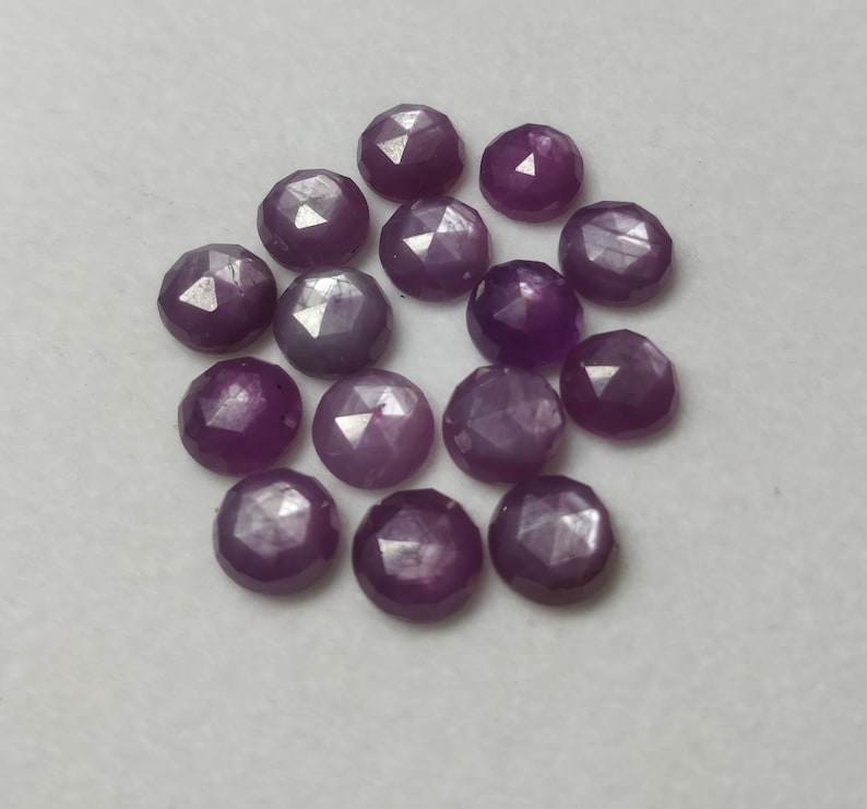 K&ouml;nnte beinhalten: Eine Gruppe von 15 violetten facettierten Edelsteinen, jeder mit einem sternf&ouml;rmigen Muster, liegt auf einer wei&szlig;en Oberfl&auml;che.