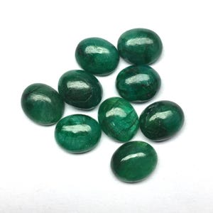 May include: Nine oval-shaped emerald gemstones, polished to a high sheen. The stones are a deep green hue and have a smooth, glossy surface. They are displayed against a white backdrop.