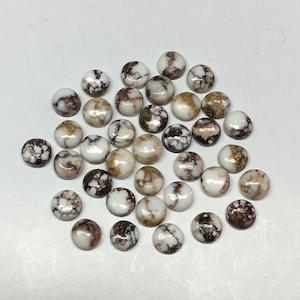 May include: A collection of round, white and brown gemstones with a marbled appearance. The stones vary in color, with some displaying more brown and others more white. The gemstones are smooth and polished, and are scattered on a white surface.