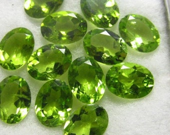 Pode incluir: Uma coleção de pedras preciosas de peridoto com corte oval. As gemas são de um verde vibrante e translúcido com múltiplas facetas, refletindo a luz. As pedras estão dispostas sobre uma superfície branca, mostrando sua clareza e cor.