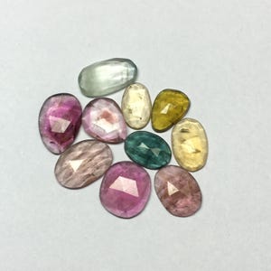 Peut inclure: Une collection de pierres pr&eacute;cieuses facett&eacute;es de diff&eacute;rentes couleurs, dont le rose, le vert, le jaune et le transparent. Les pierres pr&eacute;cieuses sont de forme ovale et en forme de goutte, avec une surface polie et r&eacute;fl&eacute;chissante. Les pierres sont dispos&eacute;es sur une surface blanche.