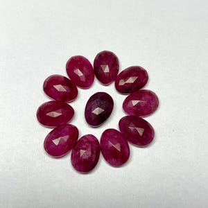 May include: Eleven oval-cut gemstones in shades of red and pink are arranged in a circle. The gemstones have multiple facets, with a darker red stone at the centre. The gemstones are displayed on a white surface.