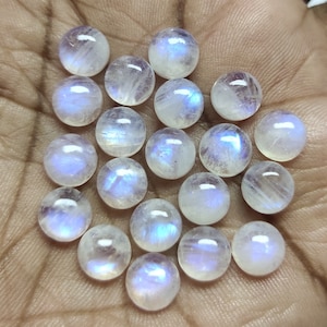 Op de afbeelding: Een verzameling van 20 ronde, witte maansteen cabochons met iriserende blauwe en paarse flitsen. De stenen zijn glad en gepolijst en zijn in een willekeurig patroon op een hand gerangschikt.