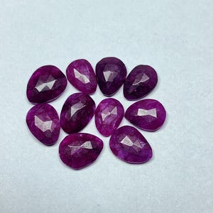 May include: Nine faceted, teardrop-shaped gemstones in shades of deep magenta are displayed. Each stone has a polished, reflective surface, creating a sparkling effect. The gems are arranged on a white surface, showcasing their colour.