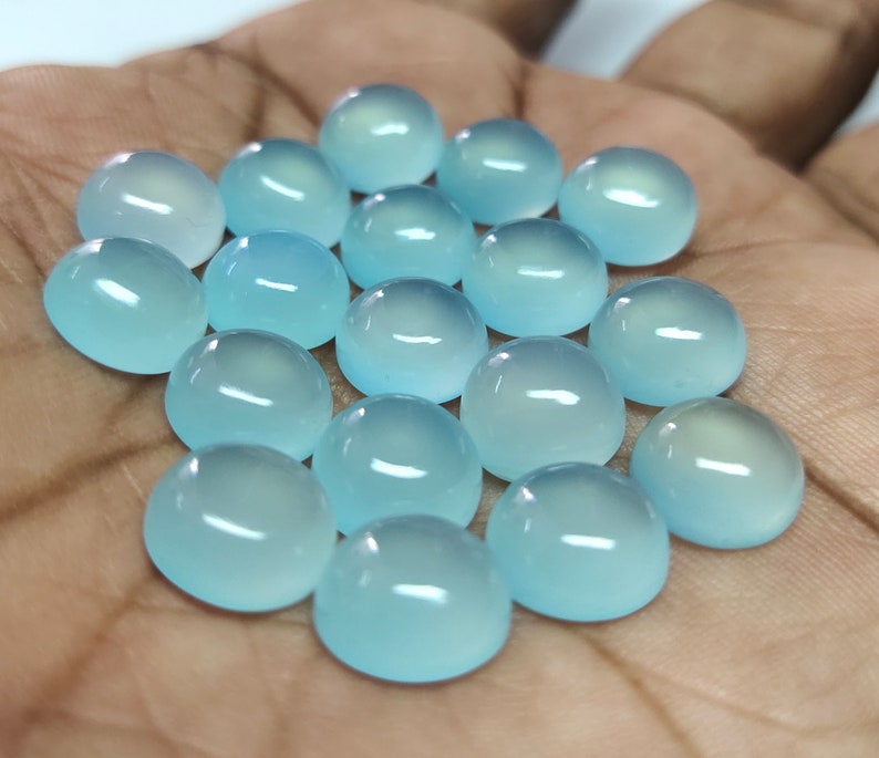 May include: A collection of 18 light blue cabochon gemstones, each with a smooth, rounded surface. The stones are arranged in a loose pile on a hand.