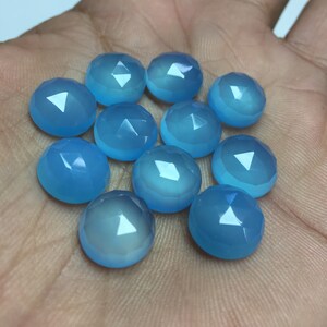 Pu&ograve; includere: Una collezione di dodici cabochon di pietre preziose blu chiaro, ognuno con una forma arrotondata sfaccettata. Le pietre sono disposte in un cerchio sciolto su una mano.