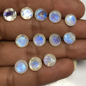 Puede incluir: Doce piedras preciosas redondas de piedra lunar blanca con destellos iridiscentes azules y blancos. Las piedras están dispuestas en dos filas en una mano.
