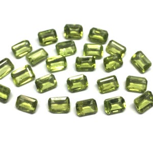 Può includere: Un gruppo di gemme rettangolari verde oliva, possibilmente peridoto, su sfondo bianco. Le gemme, lucide e sfaccettate, riflettono la luce. Le pietre, di varie dimensioni, hanno una forma rettangolare uniforme.