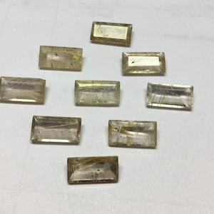 Pode incluir: Nove pedras de quartzo transparente com inclusões douradas, cortadas em forma retangular. As pedras estão dispostas em uma fileira sobre uma superfície branca.