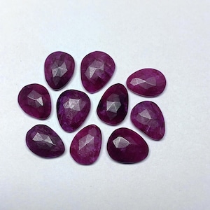 Puede incluir: Una colección de diez gemas facetadas en forma de lágrima, de varios tamaños. Las gemas son de un color magenta intenso, con múltiples facetas que capturan la luz. Están dispuestas sobre una superficie blanca.