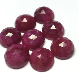May include: Eight faceted ruby gemstones, each with a deep red colour and a polished finish. The gemstones are arranged in a circular pattern on a white surface.