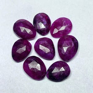 Pode incluir: Uma coleção de oito pedras preciosas facetadas, em forma oval, magenta escuro. Cada gema tem uma superfície polida com múltiplas facetas que refletem a luz. As gemas estão dispostas em um padrão circular sobre um fundo branco.