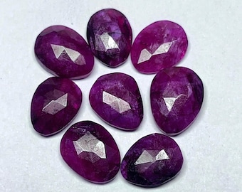 Può includere: Una collezione di otto gemme sfaccettate, di forma ovale, color magenta intenso. Ogni gemma ha una superficie lucida con molteplici sfaccettature che riflettono la luce. Le gemme sono disposte in un motivo circolare su sfondo bianco.