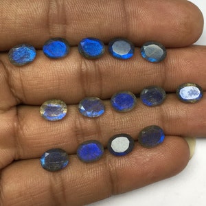 May include: A collection of oval-shaped labradorite gemstones. The stones display a mix of blue, gray, and iridescent flashes. The gemstones are arranged on a neutral background, showcasing their natural beauty and potential for jewelry making.
