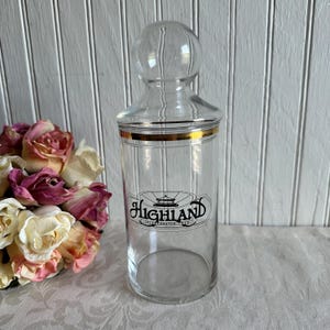 May include: A clear glass canister with a spherical lid and a gold rim around the top. The front of the cylindrical container features a black graphic logo with the text Highland Incorporated 1910, centered within an oval line design.