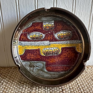 May include: Round ashtray with a metallic bronze rim and a colorful interior design. The interior features a pattern of burgundy, yellow, and white shapes. The ashtray has a small notch for resting cigarettes.
