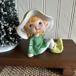 May include: A ceramic elf figurine with a green shirt, white pants, and yellow shoes. The elf has a white hat with a green brim, blonde hair, and rosy cheeks. The figurine is sitting on a brown surface.