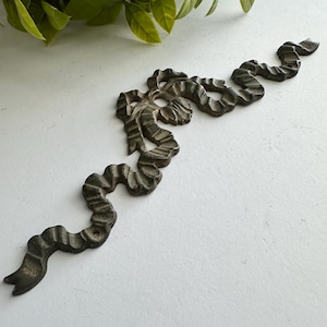May include: A dark metallic decorative wall applique shaped like a long, flowing ribbon with a central bow knot. The piece features a textured, weathered finish and includes small circular holes at the ends and center for mounting purposes.
