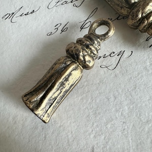 May include: A metallic gold-toned tassel charm with a weathered, antiqued finish. The charm features a rounded loop at the top for attachment, a textured, bulbous neck section, and a flared, cylindrical base with vertical grooved detailing.