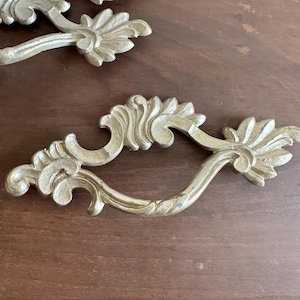 May include: Two metallic gold-toned drawer pulls featuring an ornate, curved design with leaf-like detailing at the ends. The hardware has a matte finish and a central handle section that arches outward, showcasing intricate scrollwork patterns.