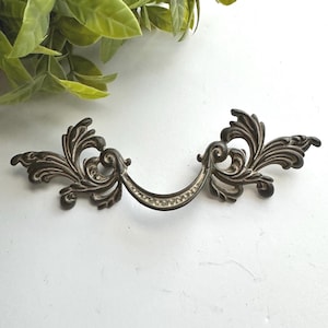 May include: A dark metal drawer pull featuring an ornate, symmetrical scrollwork design with a curved center handle. The finish has a distressed, weathered appearance with lighter highlights accentuating the intricate, raised floral-like patterns.