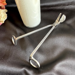 May include: Silver candle wick trimmer with a decorative etched design. The trimmer has a scissor-like shape with a circular handle and a curved blade for trimming wicks. A white candle is in the background.