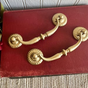 May include: Two gold-colored drawer pulls with a decorative, sunburst-like design. The pulls have a curved handle and are set against a red background. These vintage-style pulls are ideal for furniture restoration projects.