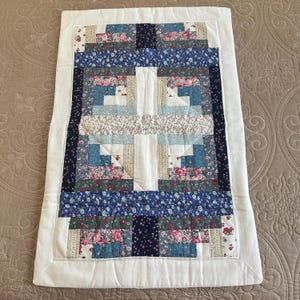 May include: A handmade quilt with a patchwork design. The quilt features a central cross pattern in white, surrounded by blocks of blue, green, and floral fabrics. A white border frames the quilt.