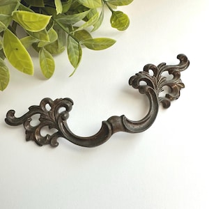 May include: A dark bronze-toned metal drawer pull featuring an ornate, curved design with intricate scrollwork and leaf-like detailing on both ends. The handle has a smooth, arched center section and a weathered, metallic finish throughout.