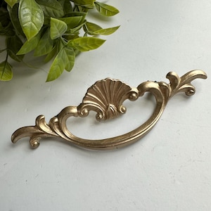 May include: A gold-colored metal drawer pull featuring an ornate, curved design with a central shell motif and symmetrical scrolling arms. The hardware has a matte finish and includes two mounting points on the back for attachment to furniture.