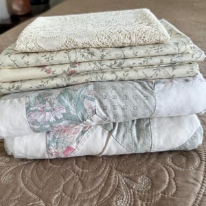 May include: A stack of folded textiles including a top layer of cream-colored lace, two middle layers featuring a light green and pink floral vine pattern on a cream background, and a bottom layer of a patchwork quilt with floral and geometric prints.
