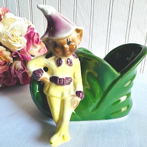 May include: A vintage ceramic planter featuring a whimsical elf figurine in a yellow outfit with purple accents. The elf stands next to a glossy green leaf-shaped planter. A bouquet of pink and cream roses adds a touch of color.