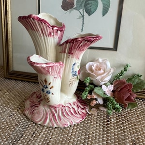 May include: A vintage ceramic vase with three openings, painted in pink, white, and blue with floral designs. The vase has a scalloped rim and a textured base.