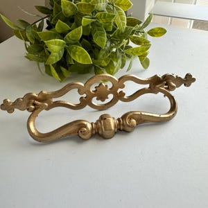 May include: A gold-colored metal drawer pull featuring an ornate, symmetrical design with a central circular motif and a hinged, curved handle. The hardware has a matte finish with subtle signs of wear, showcasing intricate scrollwork details.