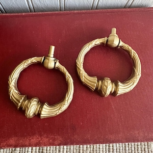 May include: Two antique brass ring pulls with ornate detailing. Each pull features a circular ring with a textured, rope-like design and decorative accents. The pulls are mounted on a red surface.