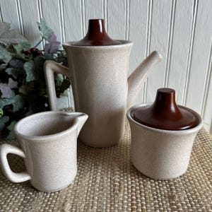 May include: A vintage coffee set featuring a speckled beige finish with dark brown accents. The set includes a coffee pot, a creamer, and a sugar bowl with a lid. The coffee pot has a tall, slender design.