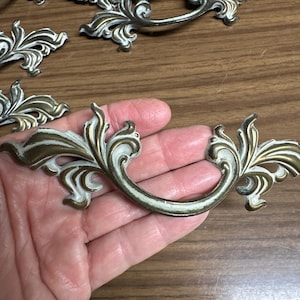 May include: A collection of metal drawer pulls featuring an ornate, curved design with a distressed finish. The hardware displays a combination of metallic gold tones and white paint, showcasing intricate scrollwork and leaf-like detailing.