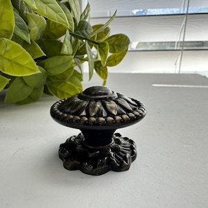 May include: A round cabinet knob with a dark bronze finish, featuring a raised petal or leaf pattern radiating from the center, a beaded outer edge, and a decorative base plate with a scalloped design that matches the top of the knob.