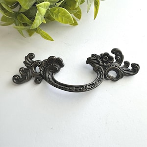 May include: A dark bronze-toned metal drawer pull featuring an ornate, curved handle with intricate scrollwork and floral detailing on both ends. The hardware has a matte finish and a symmetrical design with elaborate, sweeping decorative flourishes.