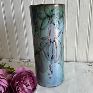 May include: A tall, cylindrical ceramic vase with a floral design. The vase features a gradient of blue and green hues, with lavender and brown floral accents. The rim is a darker brown. The vase is approximately 10 inches tall.
