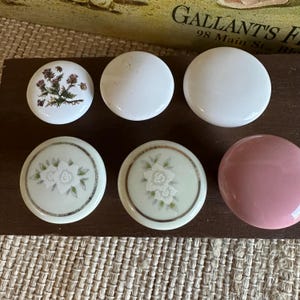 May include: A collection of ceramic cabinet knobs in various designs. The knobs include white, pink, and cream-colored options, some with floral and botanical patterns. The knobs are round and have a glossy finish, perfect for adding a decorative touch to furniture.