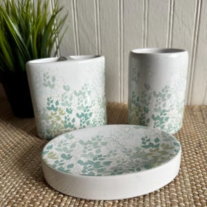May include: A white ceramic bathroom set featuring a soap dish and two cylindrical containers. The set is decorated with a pattern of green and blue-green leaves. The items are arranged on a woven mat.